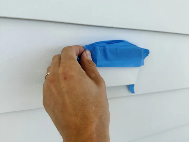 A hand pressing painter’s tape over an exterior electrical receptacle on white siding, sealing it before a soft wash begins