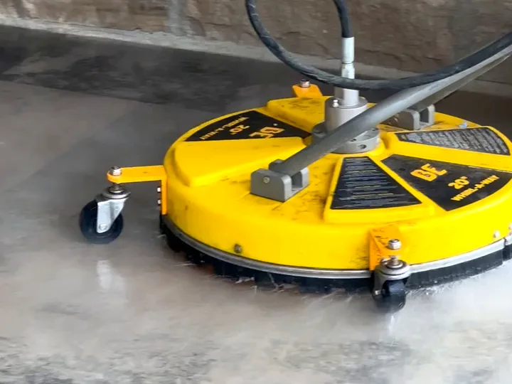 A surface cleaner mid-pass on concrete, water spraying out beneath the yellow housing — the tool that cleans a driveway evenly instead of in stripes